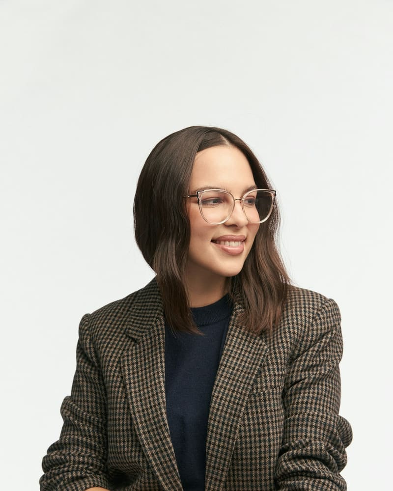 Mulher sorrindo, de óculos de grau com armação transparente e blazer xadrez